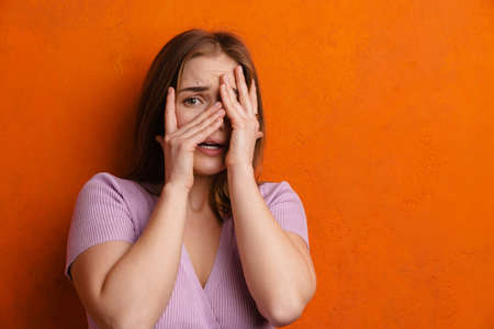 Young ginger scared woman looking at camera while covering her face isolated over orange wall