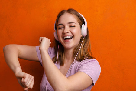 Joyful woman dancing while listening music with headphones isolated over orange wallの写真素材