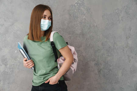 Positive young teenager student wearing protective medical mask, holding textbooks isolated over gray, looking awayの写真素材