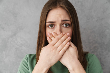 Close up of a scared shocked blonde young woman gesturing over gray wall, covers mouth