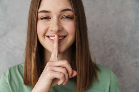 Close up of a happy blonde young woman gesturing over gray wall, showing silence gestureの写真素材
