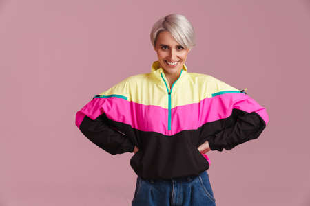 White-haired woman wearing multicolored jacket smiling at camera isolated over purple wall