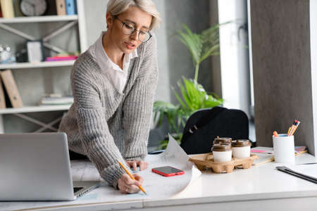 White-haired serious woman writing down notes while working with laptop in officeの写真素材