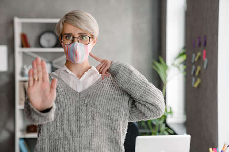White-haired woman pointing finger at her face mask while showing stop gesture in office