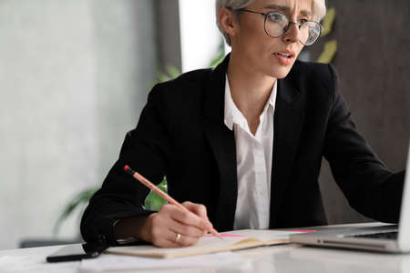 White-haired puzzled woman writing down notes while working with laptop in office
