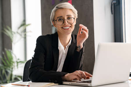 White-haired joyful woman working with laptop while sitting at table in office