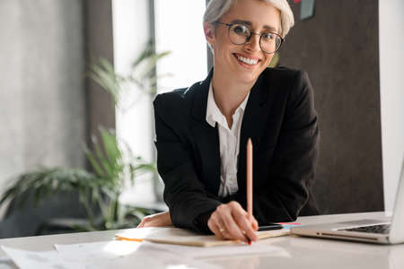 White-haired joyful woman working with laptop while sitting at table in officeの写真素材
