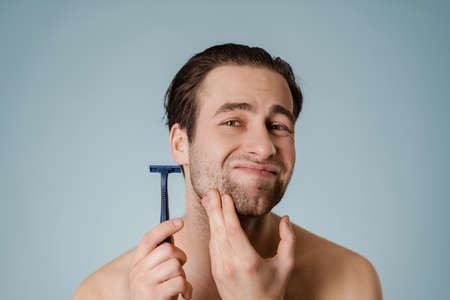 Close up portrait of a young shirtless man shaving with a razor isolated over blue backgroundの写真素材