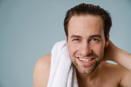 Shirtless white man smiling at camera while posing with towel isolated over blue wall