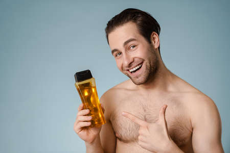 Close up of a smiling handsome young nude man presenting shampoo bottle isolated over blue backgroundの写真素材