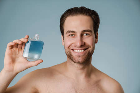Shirtless white man smiling while showing cologne isolated over blue wallの写真素材