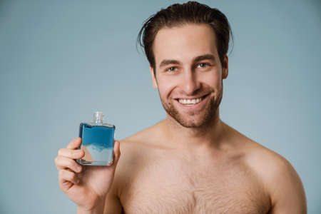 Shirtless white man smiling while showing cologne isolated over blue wallの写真素材