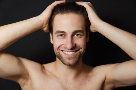 Shirtless white man smiling while holding his head isolated over black wallの写真素材