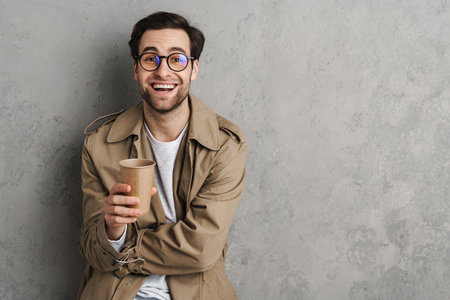 Handsome man wearing stylish coat posing isolated over gray background, drinking coffeeの写真素材