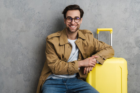 Happy handsome guy smiling while posing with suitcase isolated over grey wallの写真素材