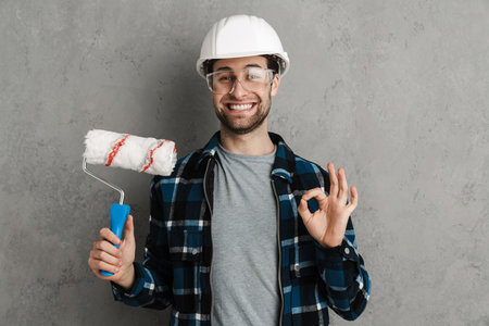 Smiling man in protective helmet hardhat holding paint brush isolated on gray wall background, showing okの写真素材