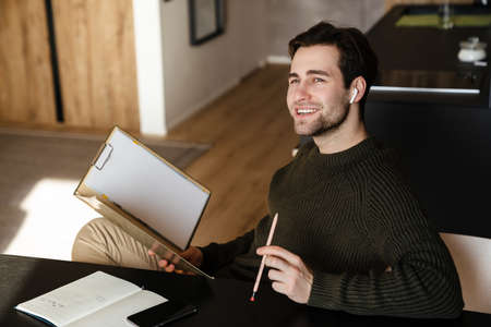 Smiling brunette man taking notes while sitting at tbe table at homeの写真素材