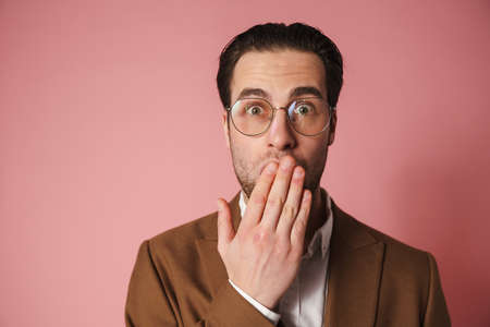 Shocked brunette man in jacket looking at camera over pink wall background, cover mouth