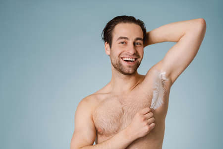 Attractive young smiling nude man touching his body with a feather isolated over blue backgroundの写真素材