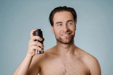 Handsome smiling nude man spraying deodorant isolated over blue backgroundの写真素材