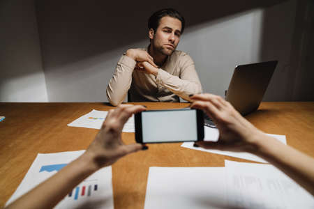 Serious brunette entrepreneur sitting in the office during interview with a boss, holding blank screen mobile phoneの写真素材