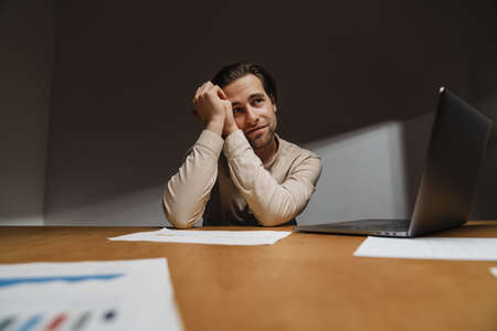 Pensive brunette businessman working on laptop computer in dark officeの写真素材