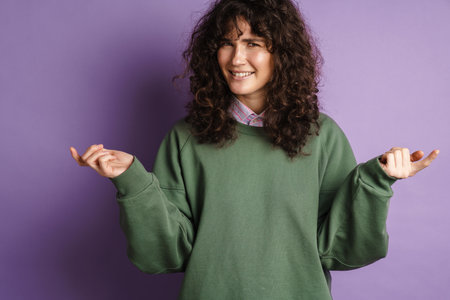 Happy puzzled curly girl smiling and gesturing with hands isolated over purple backgroundの写真素材