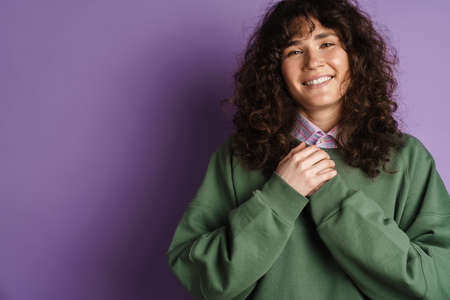 Happy brunette curly girl smiling and holding her hand at chest isolated over purple background