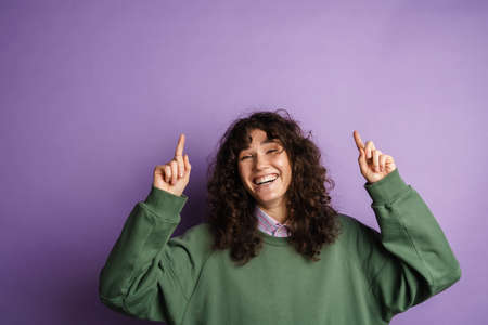Happy beautiful curly girl smiling and pointing her fingers upward isolated over purple background