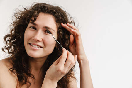 Half-naked curly woman smiling and using brow brush isolated over white background