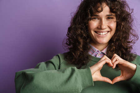 Happy beautiful curly girl smiling and gesturing heart sign with fingers isolated over purple...