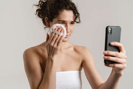 European curly woman using powder sponge and cellphone isolated over white backgroundの写真素材
