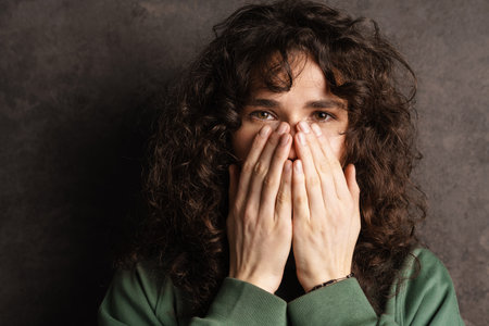 Young brunette curly girl covering her mouth with hands isolated over black wall