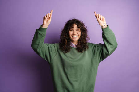 Happy beautiful curly girl smiling and pointing her fingers upward isolated over purple background