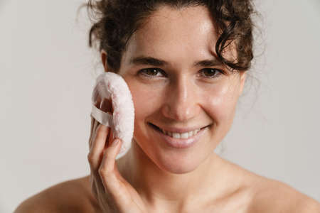 Half-naked curly woman smiling and using powder sponge isolated over white backgroundの写真素材