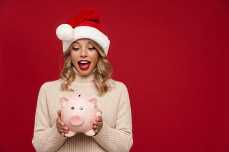 Excited pretty young woman wearing santa hat holding piggybank isolated over red backgroundの写真素材
