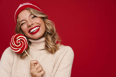 Beautiful happy girl in winter hat smiling while posing with lollipop isolated over red backgroundの写真素材