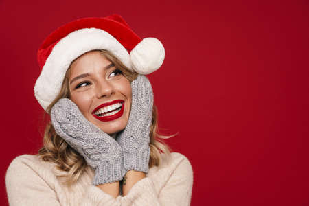 Nice happy girl in Santa Claus hat and knitted mittens posing isolated over red backgroundの写真素材