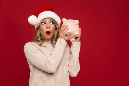 Excited pretty young woman wearing santa hat holding piggybank isolated over red backgroundの写真素材