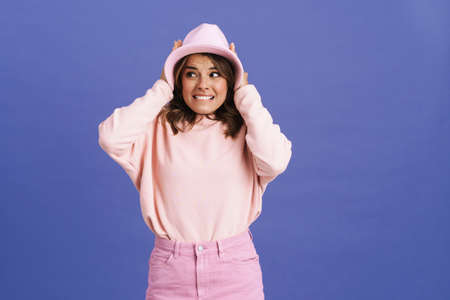 Scared charming girl in hat grabbing her head and looking aside isolated over purple backgroundの写真素材
