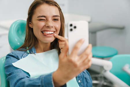 European young woman showing her tongue while taking selfie on cellphone at dental clinicの写真素材