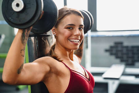 Image of happy positive young woman fitness coach making sport exercise for arms with dumbbells in gymの写真素材
