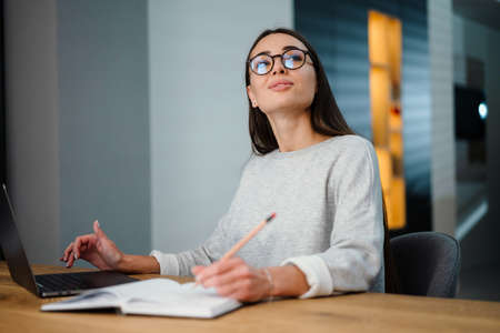 Happy young woman writing down notes while working with laptop at homeの写真素材