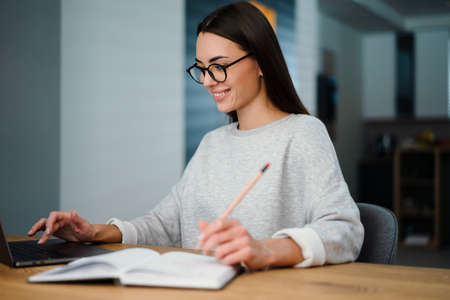 Happy young woman writing down notes while working with laptop at homeの写真素材