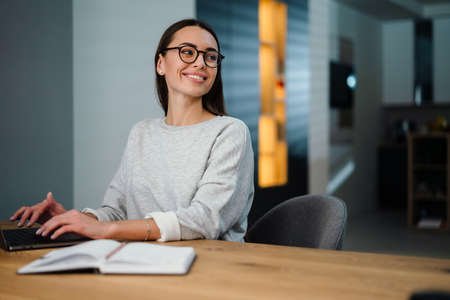Happy young woman in eyeglasses smiling while working with laptop at homeの写真素材