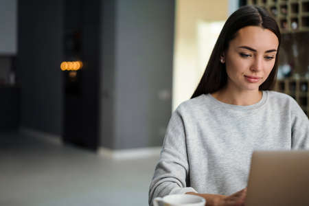 Focused young woman working with laptop and drinking coffee at home