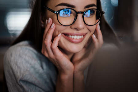 Happy young woman in eyeglasses smiling while working with laptop at homeの写真素材