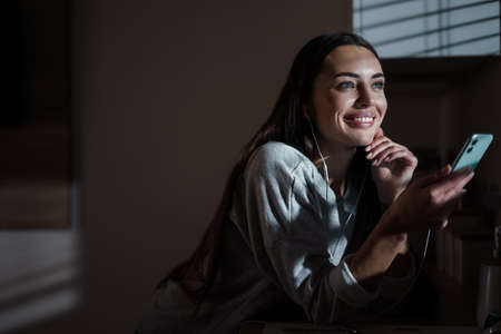 Joyful young woman in earphones smiling while using cellphone at home kitchen