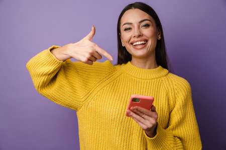 Happy young woman wearing sweater pointing finger at mobile phone over violet background