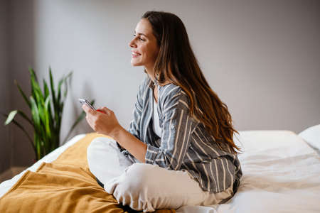 Pleased brunette woman using cellphone while sitting on bed at homeの写真素材
